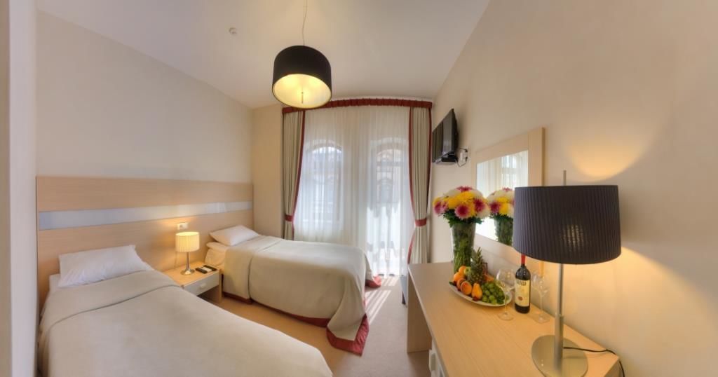 A hotel room with two beds, a desk with a lamp, a vase of flowers, and a wine bottle.