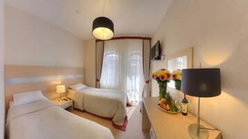A hotel room with two beds, a desk with a lamp, a vase of flowers, and a wine bottle.