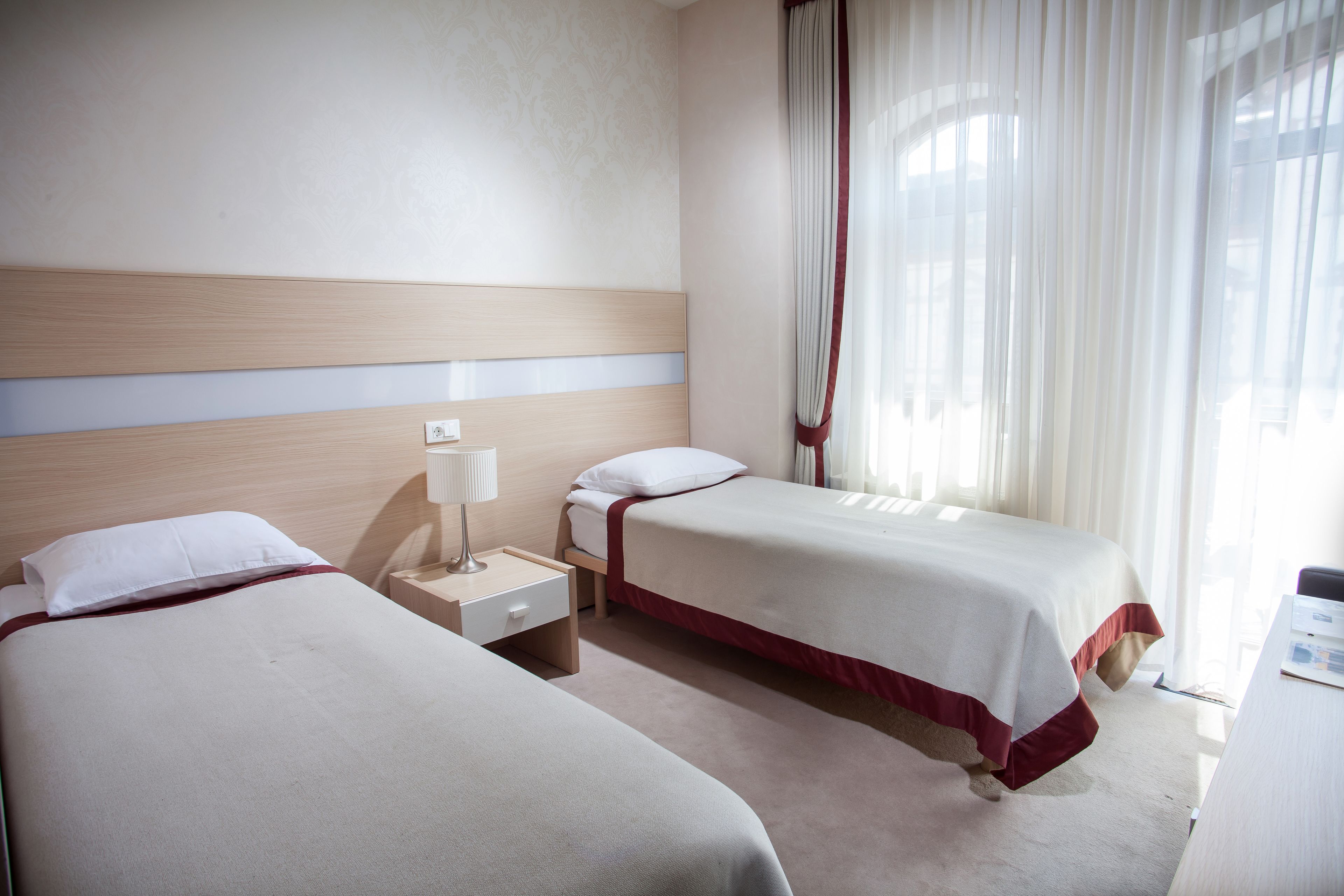 A hotel room with two beds, a nightstand, a lamp, and a window with curtains.