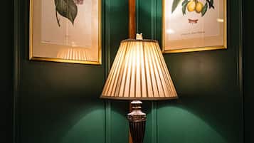 Quarto com paredes verdes, uma mesa redonda de madeira, uma luminária, duas ilustrações botânicas emolduradas, um livro e um vaso com flores.