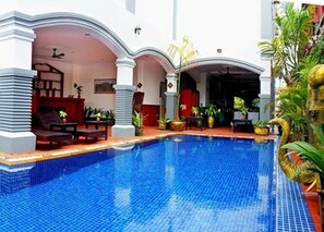 Una piscina con bordes de azulejos, rodeada de un porche cubierto con asientos y plantas en macetas.