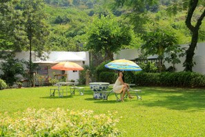 A woman sitting on a chair under a colorful umbrella in a grassy outdoor area with tables and a large decorative bowl. - Oritel Service Apartments (Mumbai)
