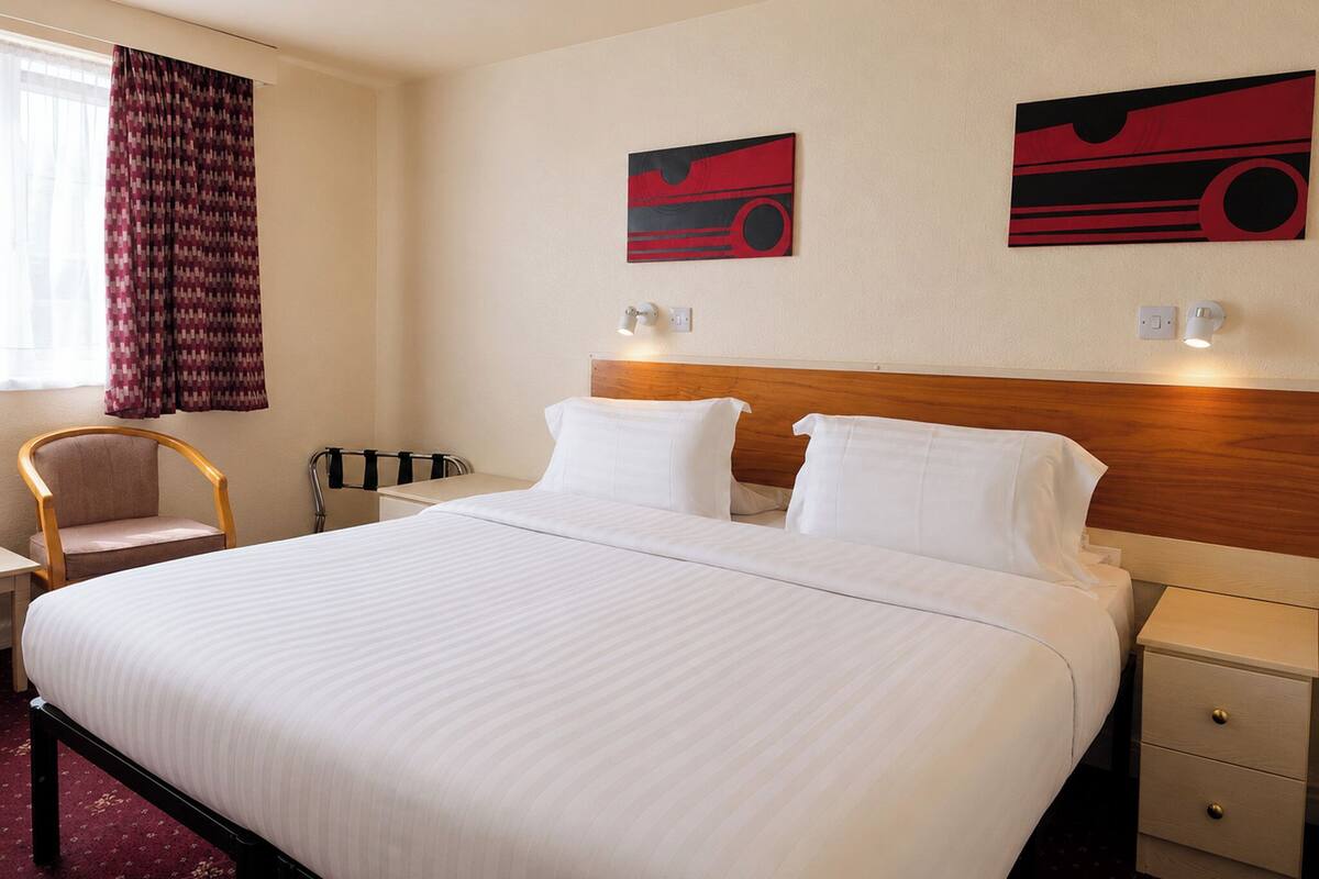 Habitación de hotel con una cama grande, cabecera de madera, dos lámparas fijadas a la pared, una silla, una mesita de noche y una ventana con cortinas.