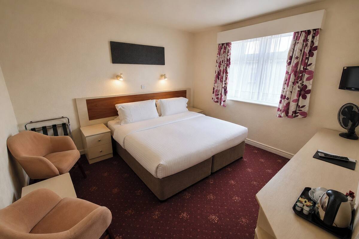 Habitación de hotel con cama, mesitas de noche, una silla, escritorio con ventilador y ventana con cortinas.