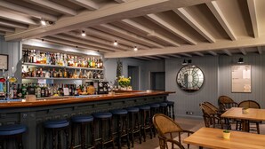 Zona de bar con barra de madera, taburetes y gran variedad de botellas. Hay sillas y una mesa con una planta encima.