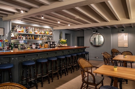 Zona de bar con barra de madera, taburetes y gran variedad de botellas. Hay sillas y una mesa con una planta encima.