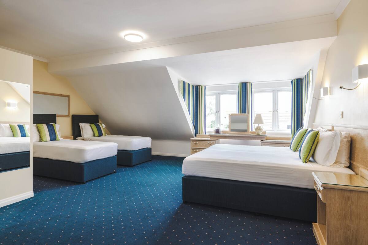 Habitación de hotel con dos camas, un escritorio, una silla y un ventanal con cortinas.