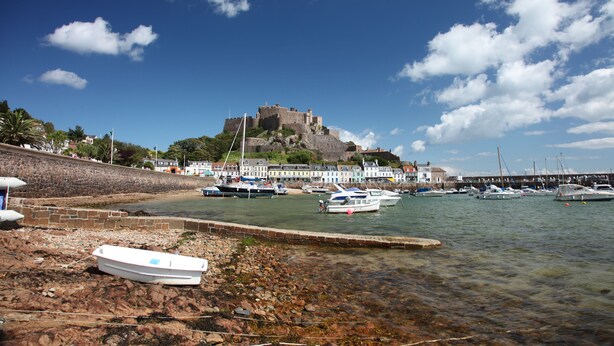 Un pueblo costero con un castillo histórico en una colina, un puerto deportivo con barcos y un litoral rocoso.