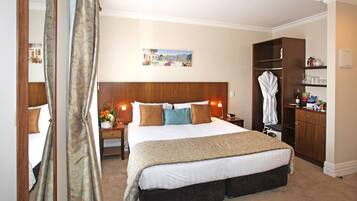 Habitación de hotel con una cama grande, cabecera de madera, mesita de noche con lámpara, bata colgada en un perchero y un mueble pequeño con artículos de aseo.