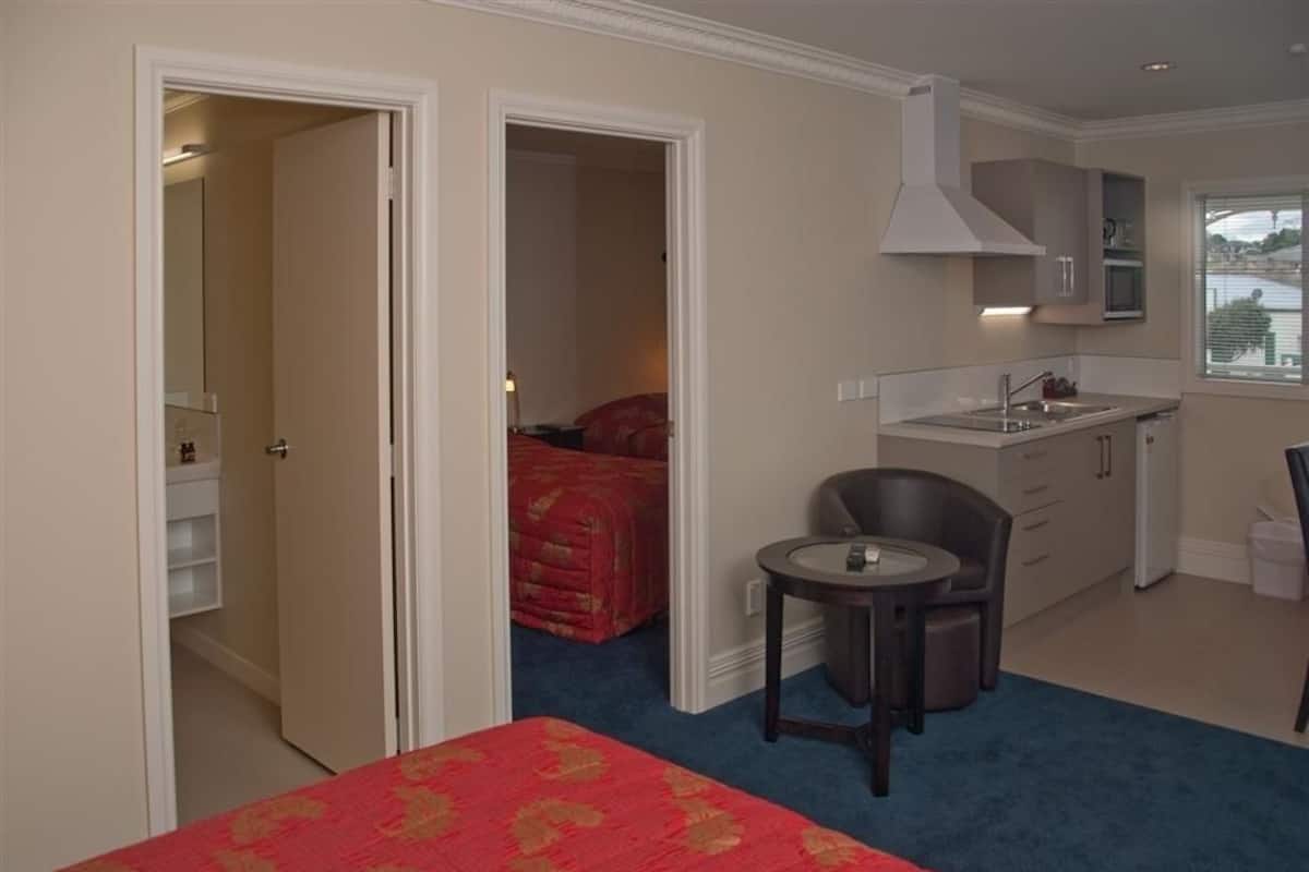 A hotel room with a bed, a small table, a chair, a kitchenette, and a bathroom visible through an open door.
