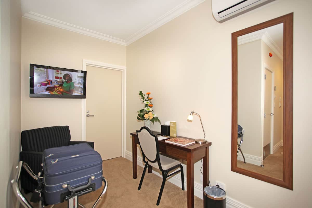 A hotel room with a desk, chair, and a television displaying a program. There is a suitcase on a chair and a trash can near the desk.