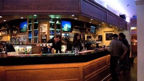 A bar area with a wooden counter, shelves with bottles, and a television screen.