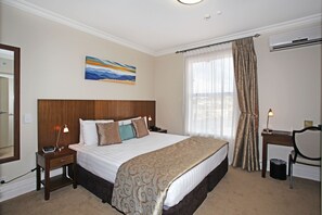 A hotel room with a large bed, a desk, a chair, a nightstand, a lamp, a painting, and a window with curtains. - Tuakau Hotel (Tuakau)