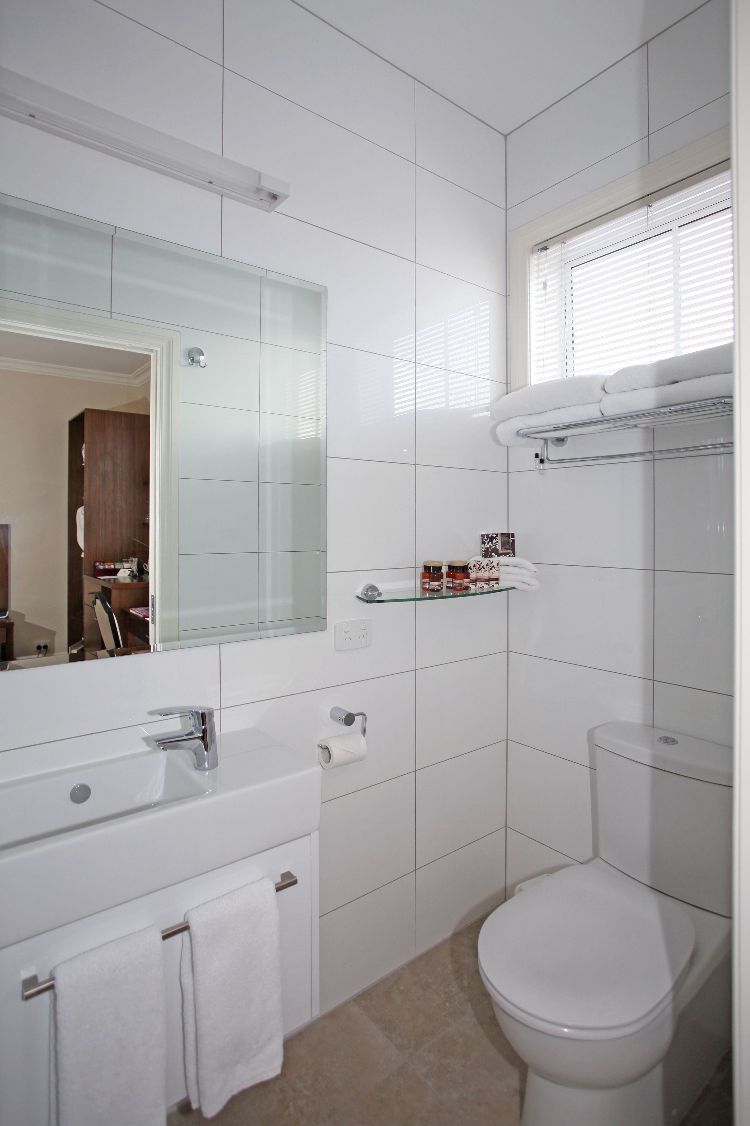 A modern bathroom with white tiles, a glass shower enclosure, a white toilet, and a vanity with a mirror and towel rack.