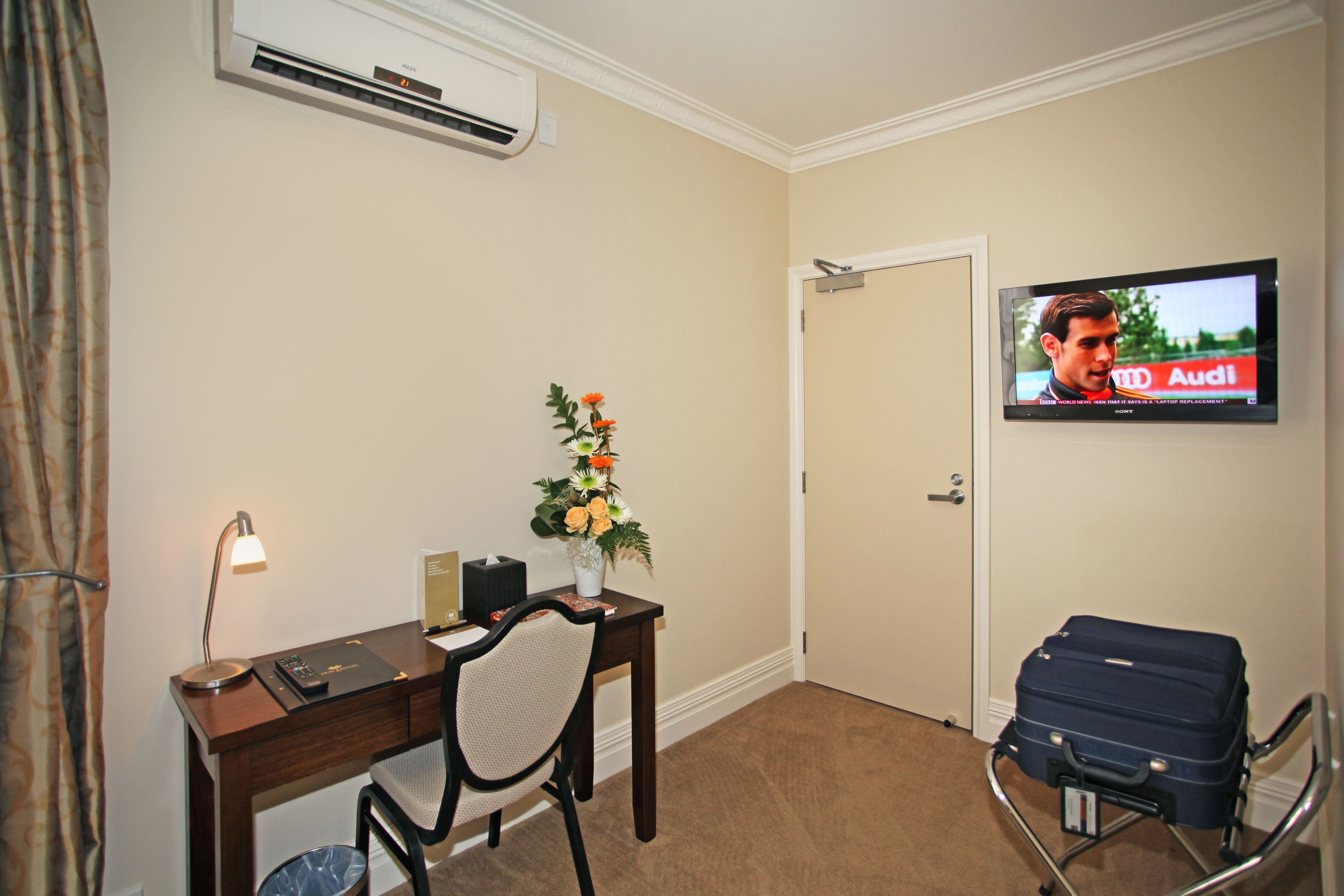 Une chambre d’hôtel équipée d’un bureau, d’une chaise, d’un climatiseur, d’une télévision et d’une valise.