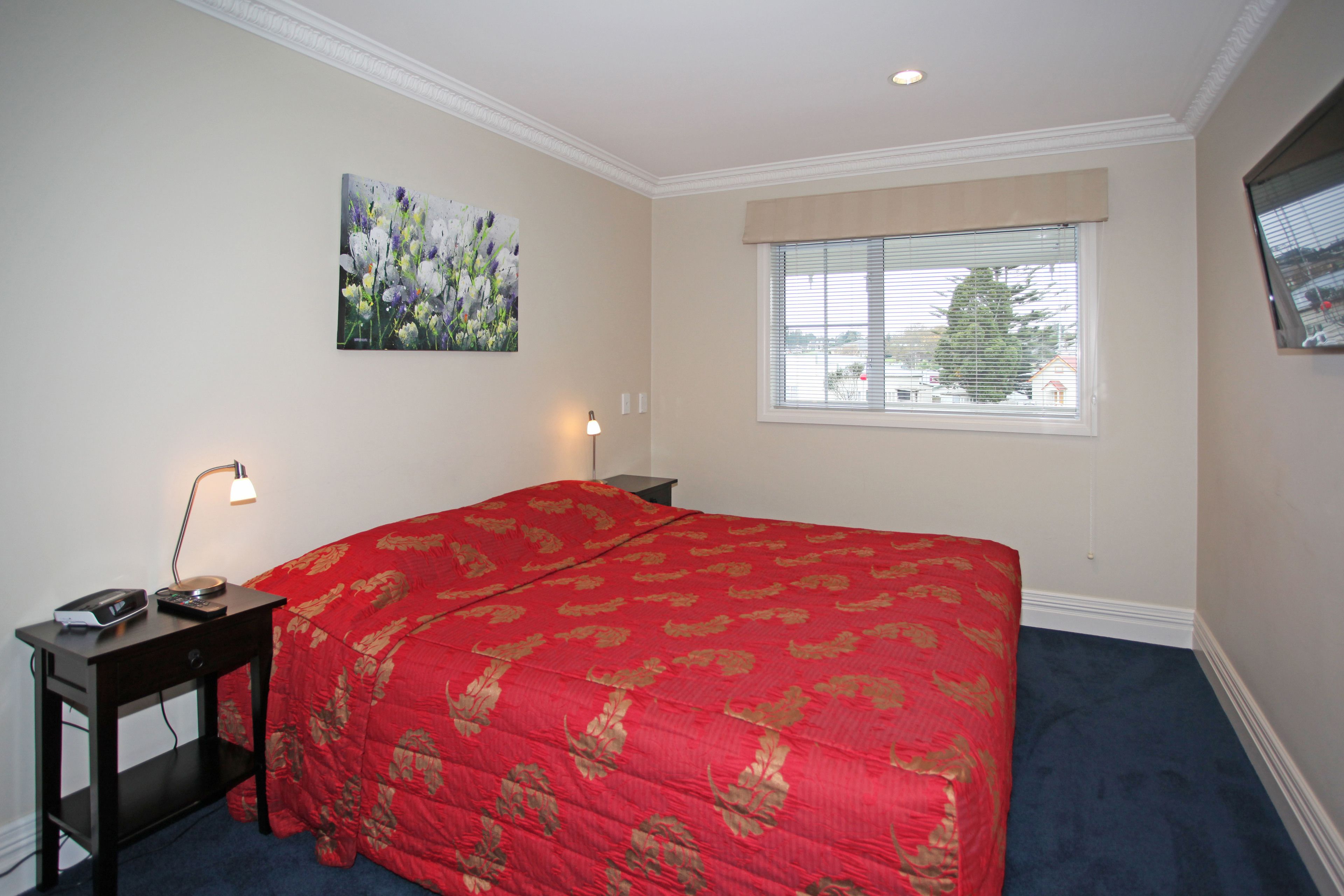 A bedroom with a large bed, a nightstand with a lamp, a painting on the wall, and a window with blinds.
