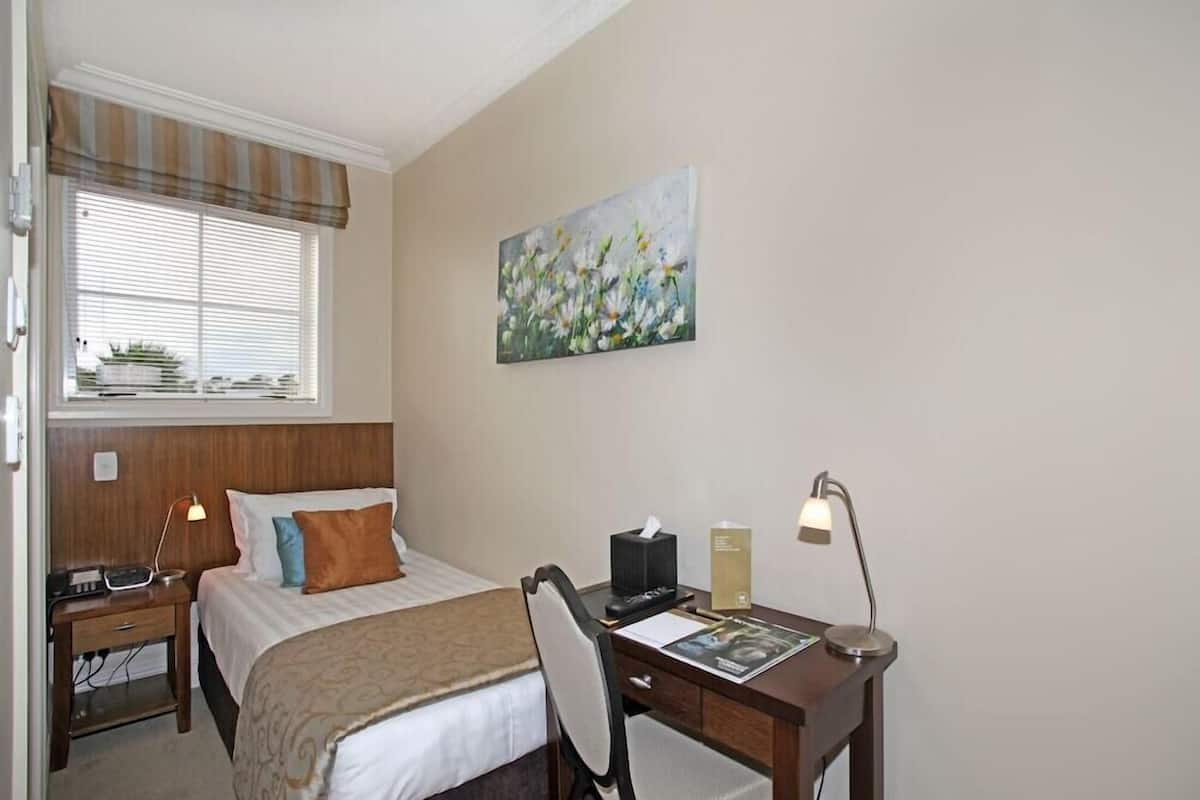 A hotel room with a bed, bedside table with lamp, desk with chair, and artwork on the wall.