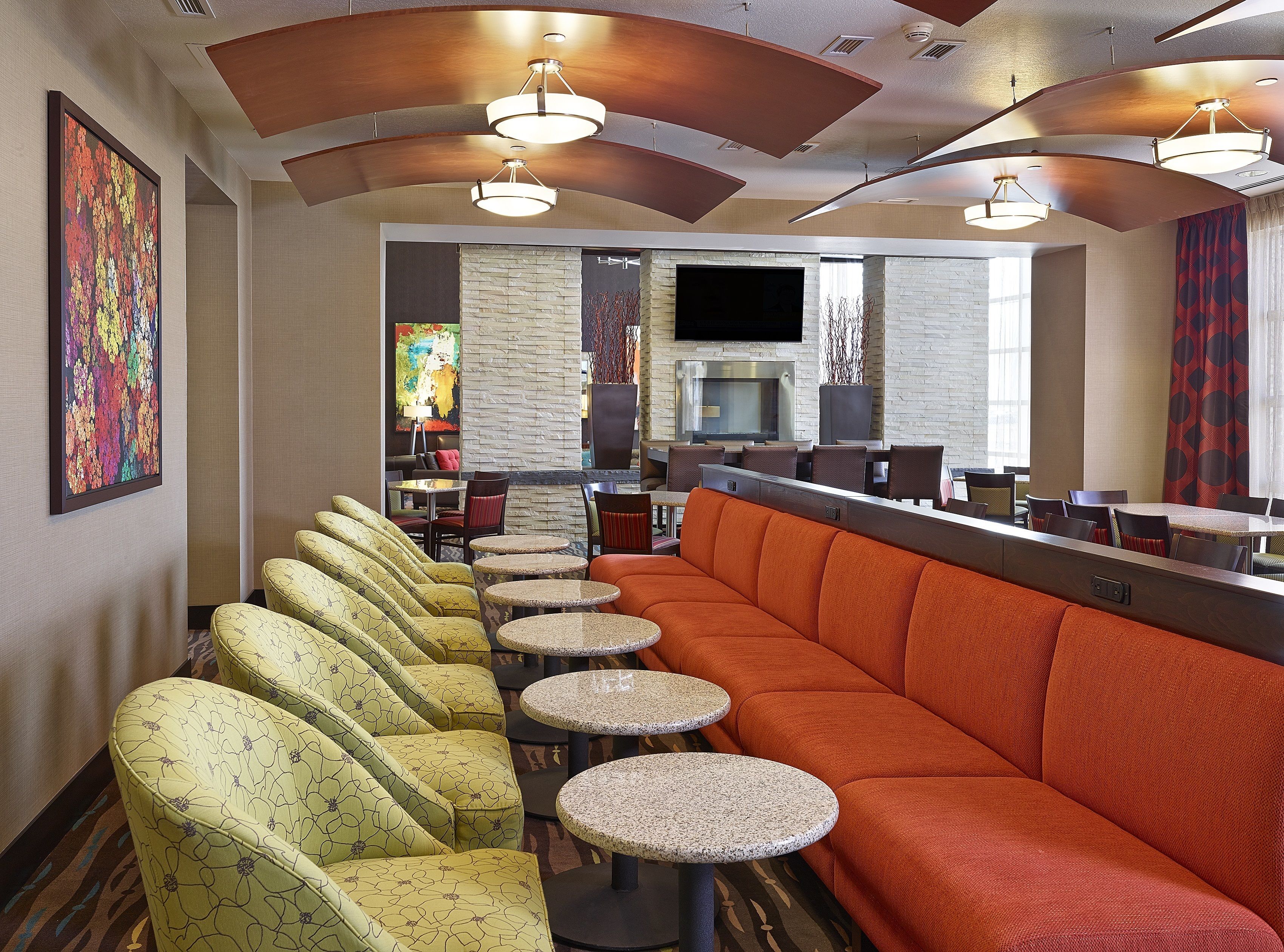 A restaurant with a long orange sofa, yellow patterned chairs, and round tables.