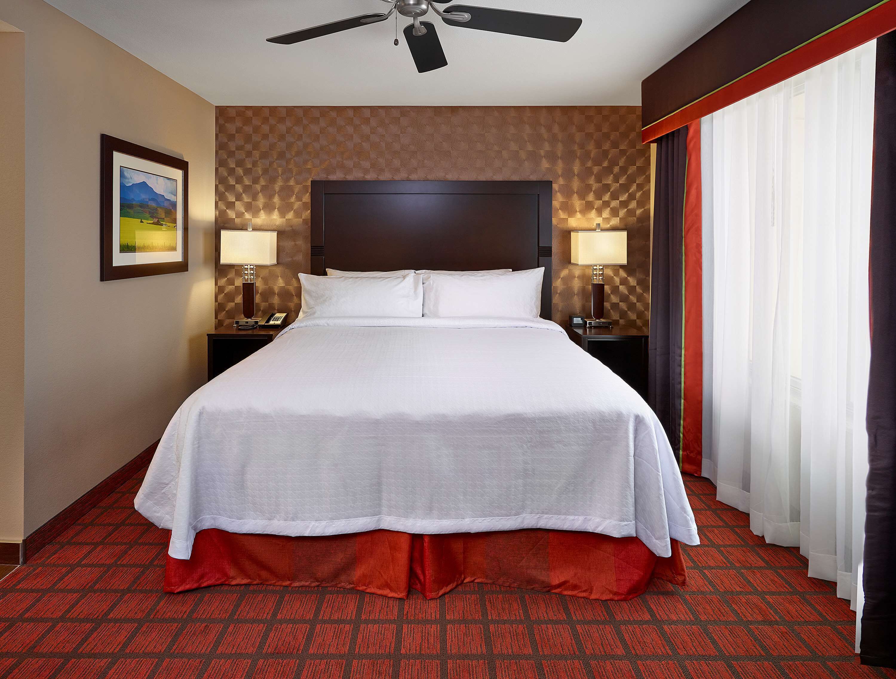 Una habitación de hotel con una cama grande, dos mesitas de noche con lámparas, un ventilador de techo, un cuadro en la pared y una ventana con cortinas.