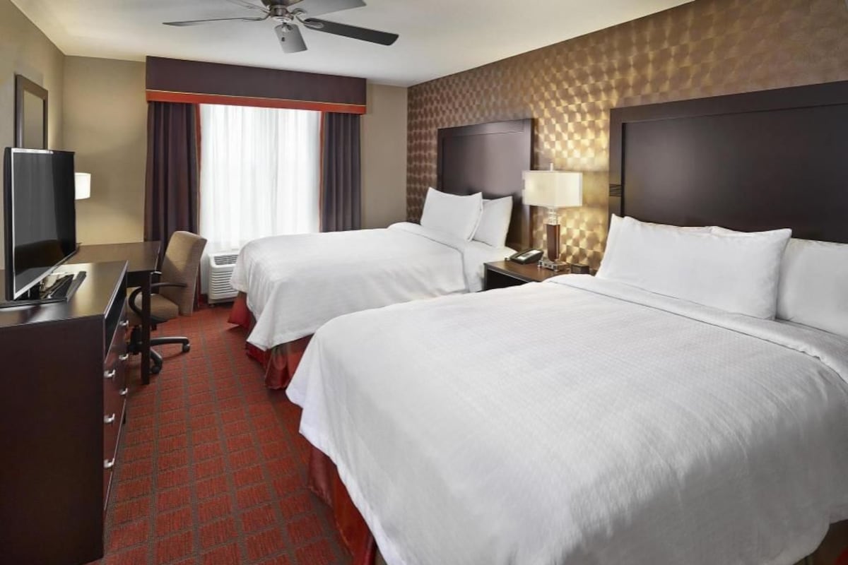 A hotel room with two beds, a desk with a TV, a chair, a desk lamp, and a ceiling fan.