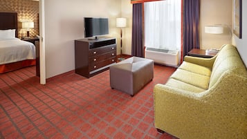Habitación de hotel con cama, televisor, cómoda, otoman, sofá y ventana con cortinas.