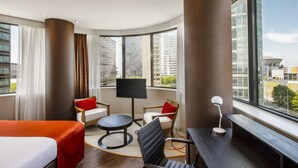 A hotel room with a large window, a bed, a desk, a chair, and a TV.