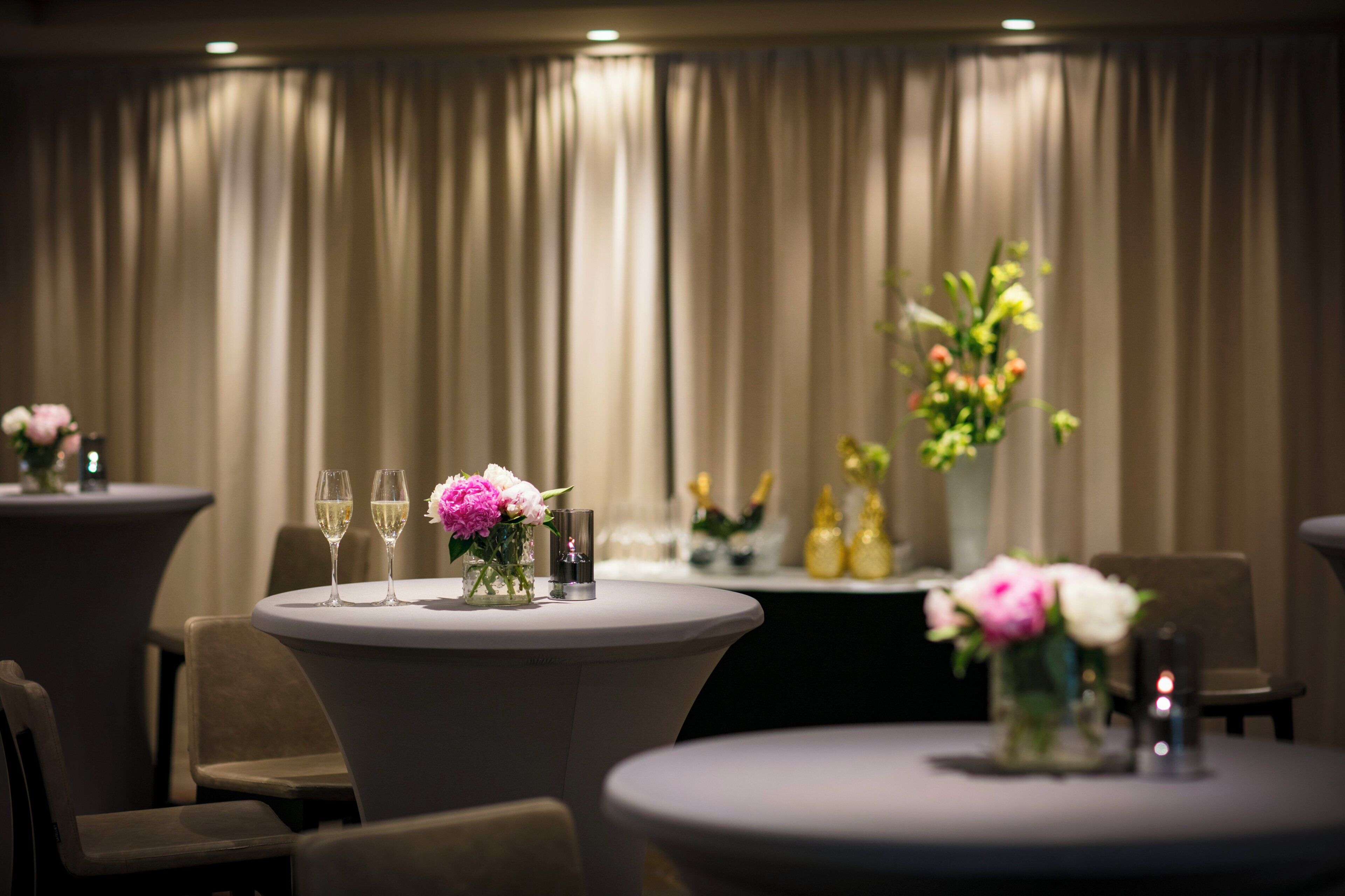 A hotel dining area with round tables, each with a vase of flowers and two glasses of wine.