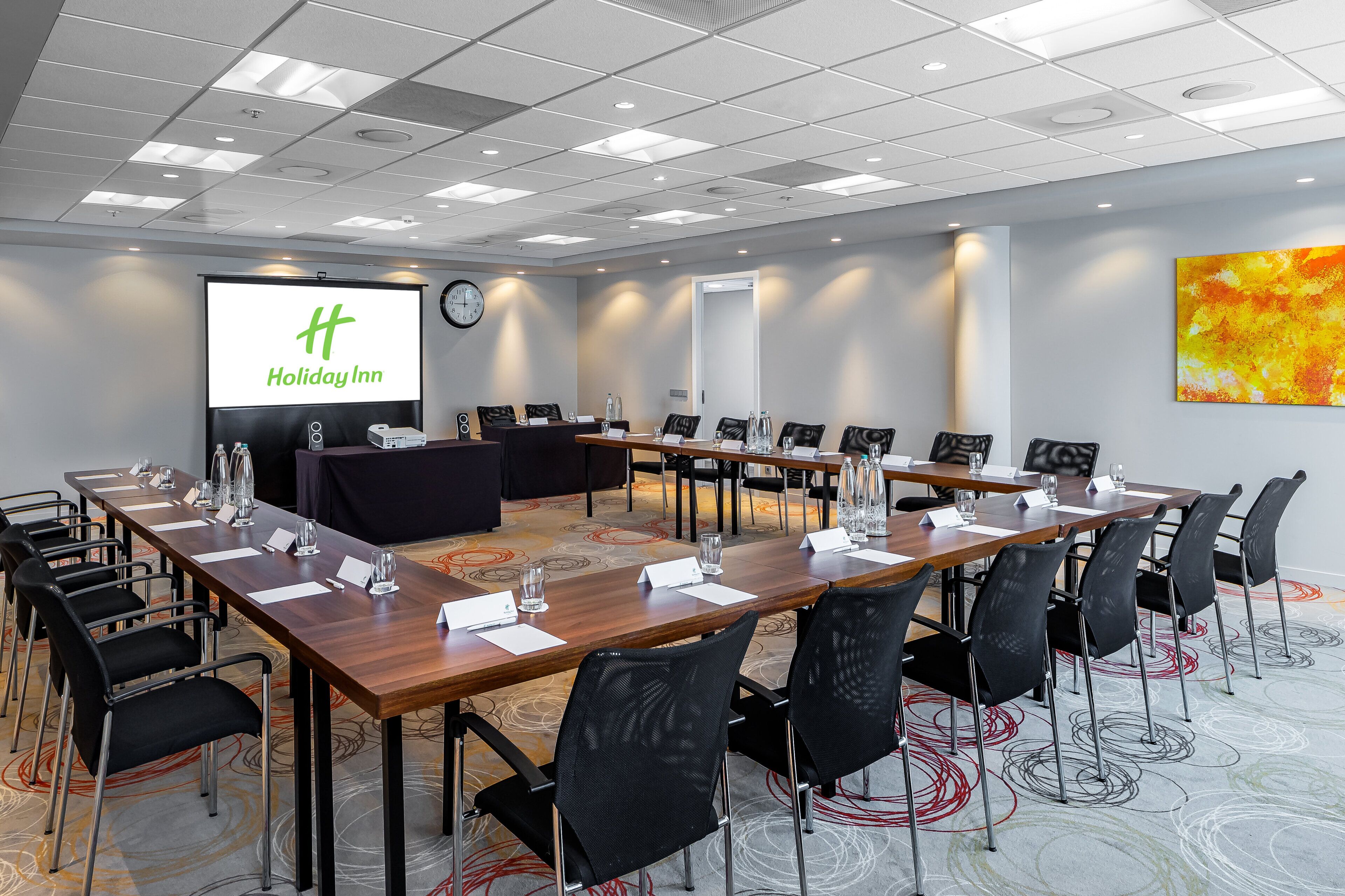 Un salón de conferencias con una pantalla grande que muestra el logotipo de Holiday Inn, varias mesas con sillas y suelo alfombrado.