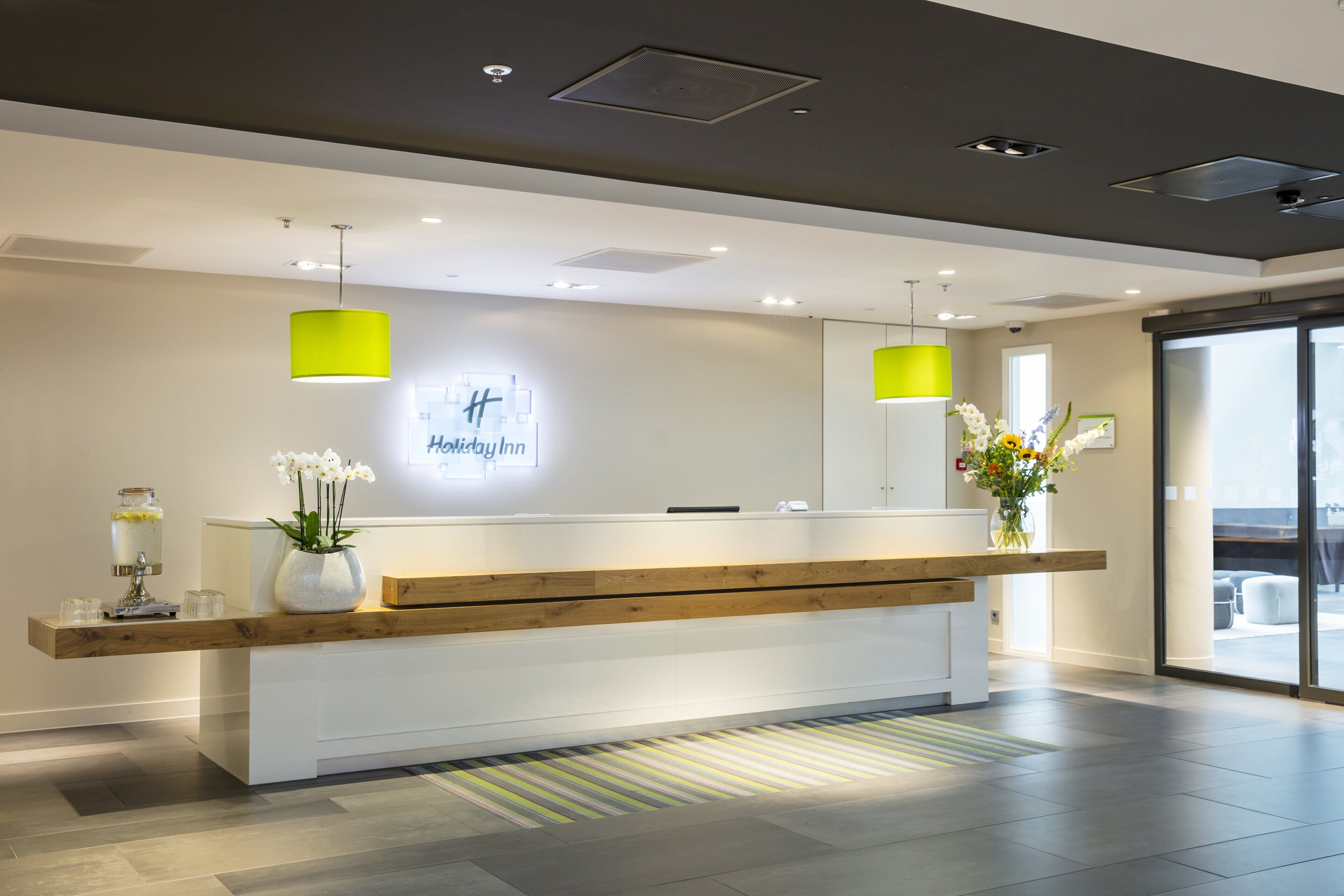 Een modern receptiebalie met een houten aanrecht, een glazen vaas met bloemen en een bord met de tekst 'Holiday Inn'.