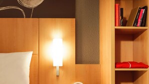A hotel room with a bed, a bedside table with a tablet and headphones, a wall-mounted lamp, and a bookshelf with books and a red towel.