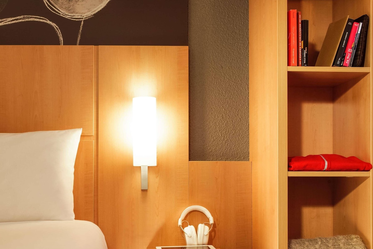 A hotel room with a bed, a bedside table with a tablet and headphones, a wall-mounted lamp, and a bookshelf with books and a red towel.