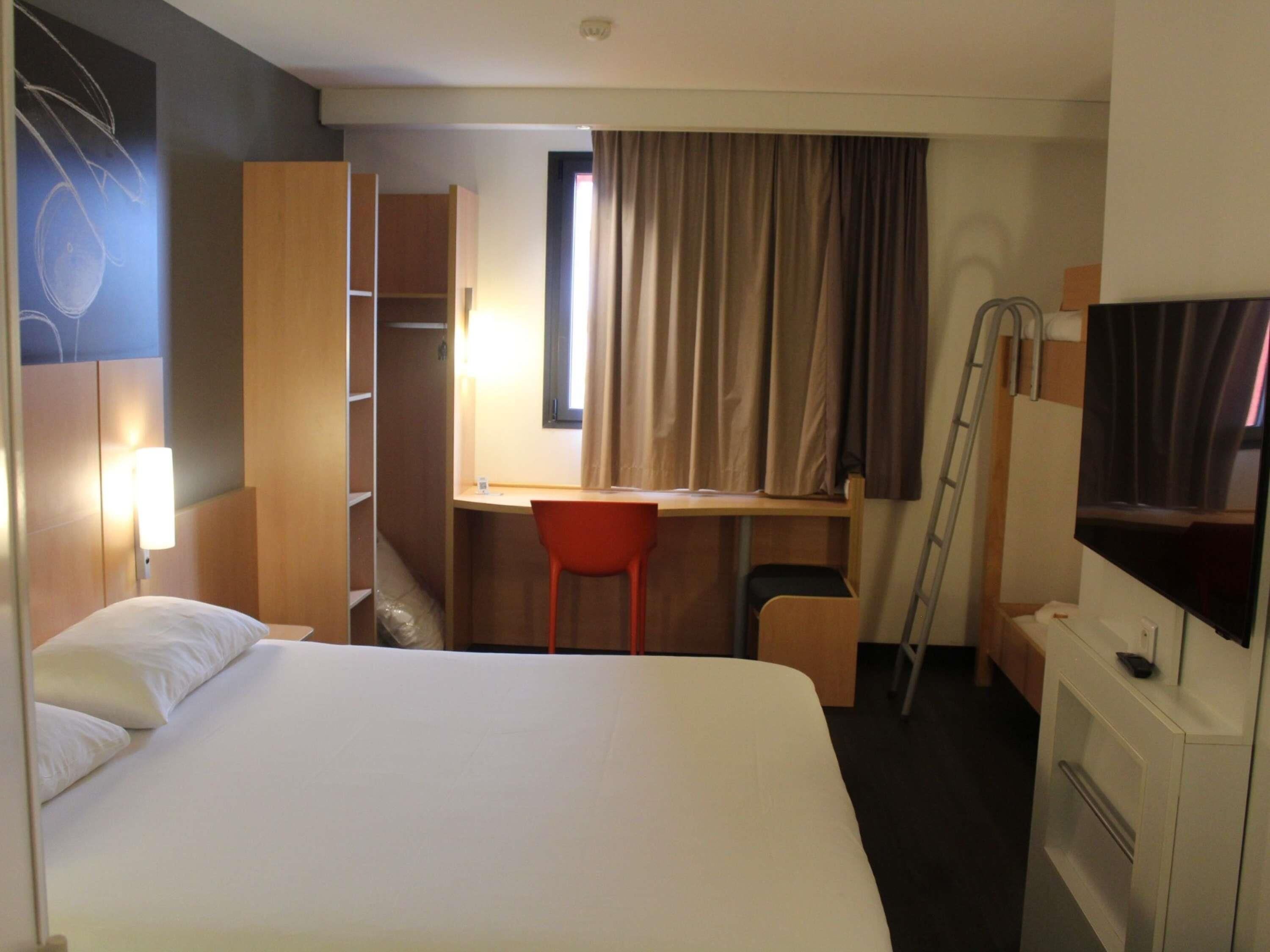 A hotel room with a bed, a desk, a chair, a wardrobe, a TV, and a window with curtains.