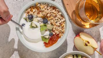 Eine Schüssel Joghurt mit Früchten und Granola, ein Glas bernsteinfarbener Flüssigkeit und ein halber Apfel auf einem Tisch.