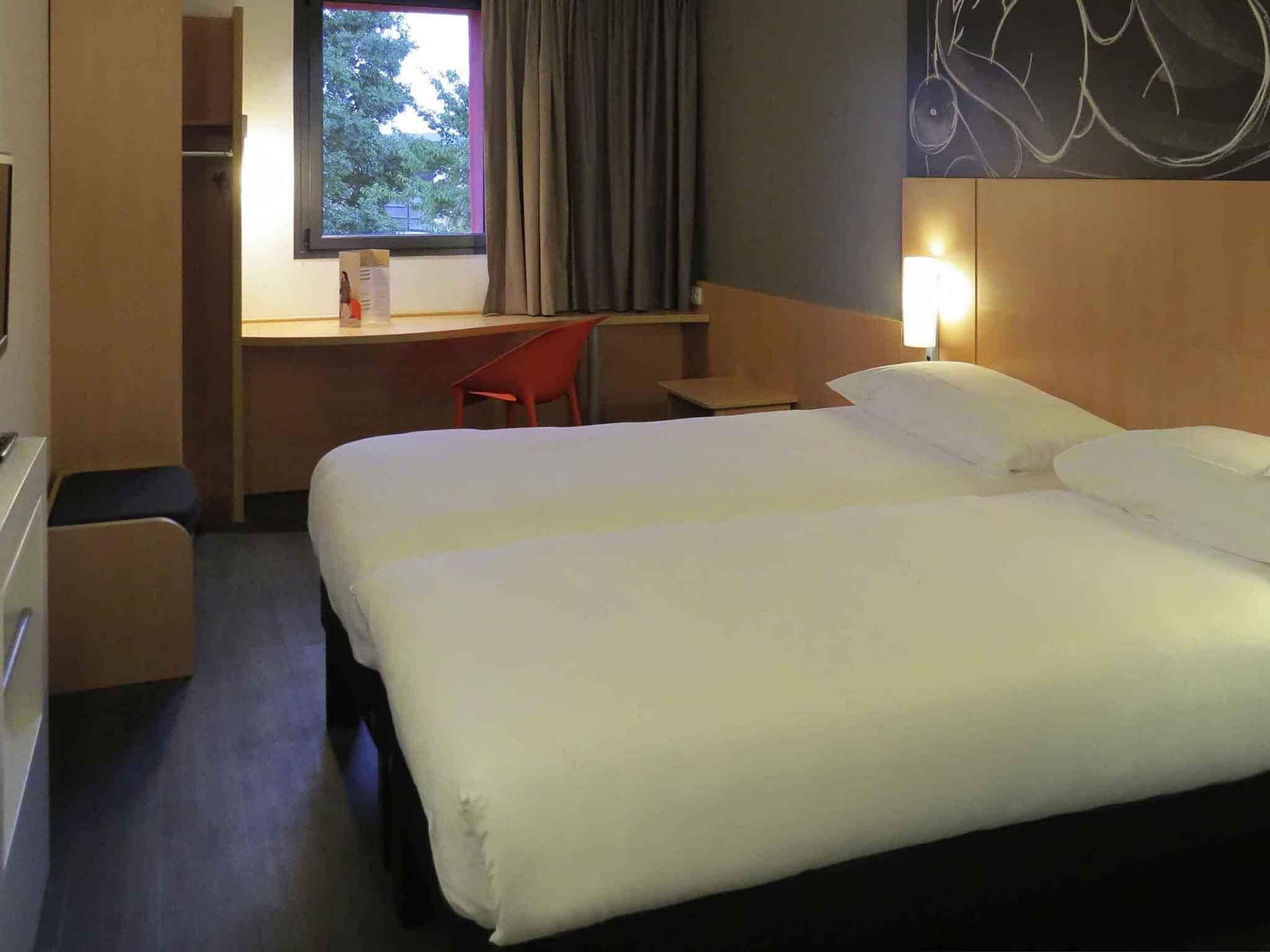 A hotel room with a double bed, a desk with a chair, a small table, and a window with curtains.