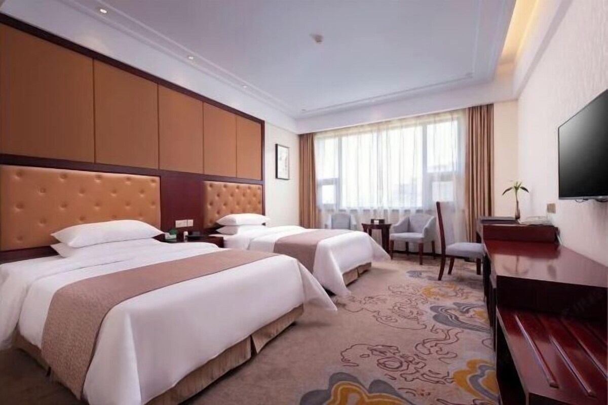 A hotel room with two beds, a flat-screen TV, a desk, and a large window with curtains.