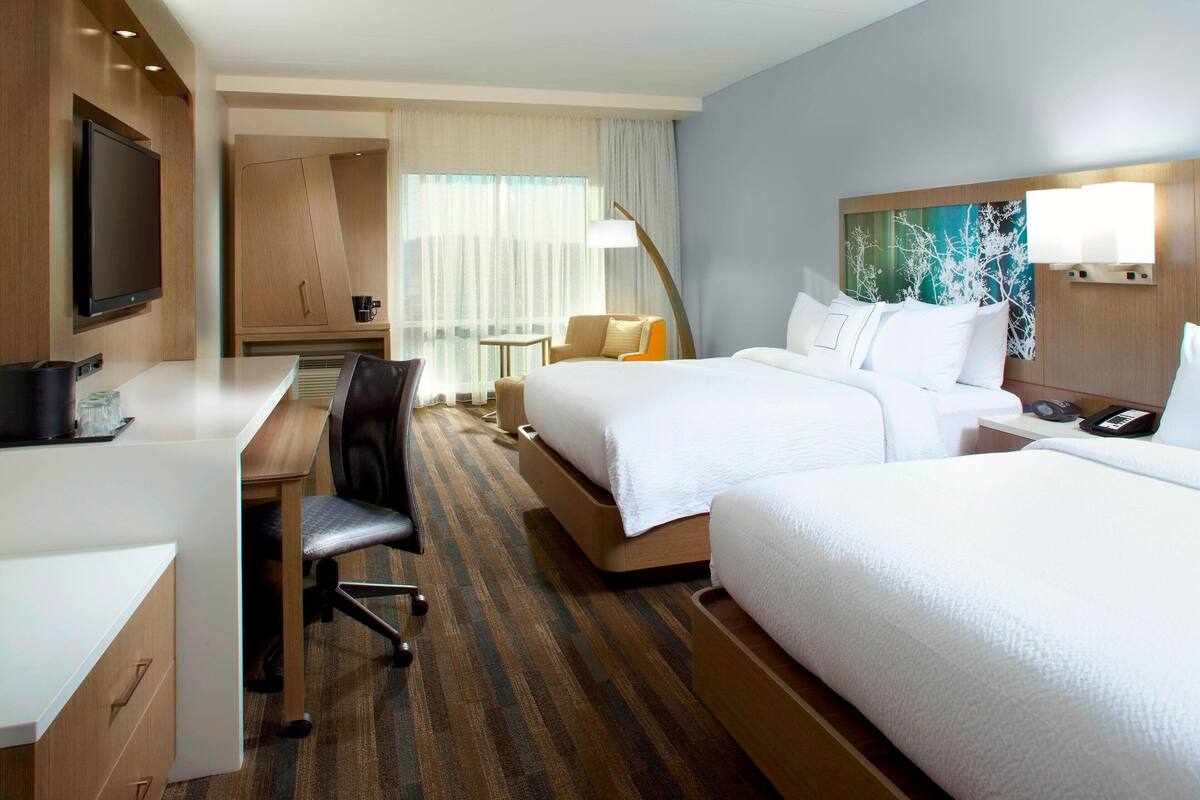 A hotel room with a desk, two beds, a TV, and a painting on the wall.