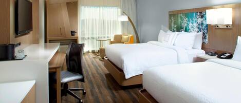 A hotel room with a desk, two beds, a TV, and a painting on the wall.