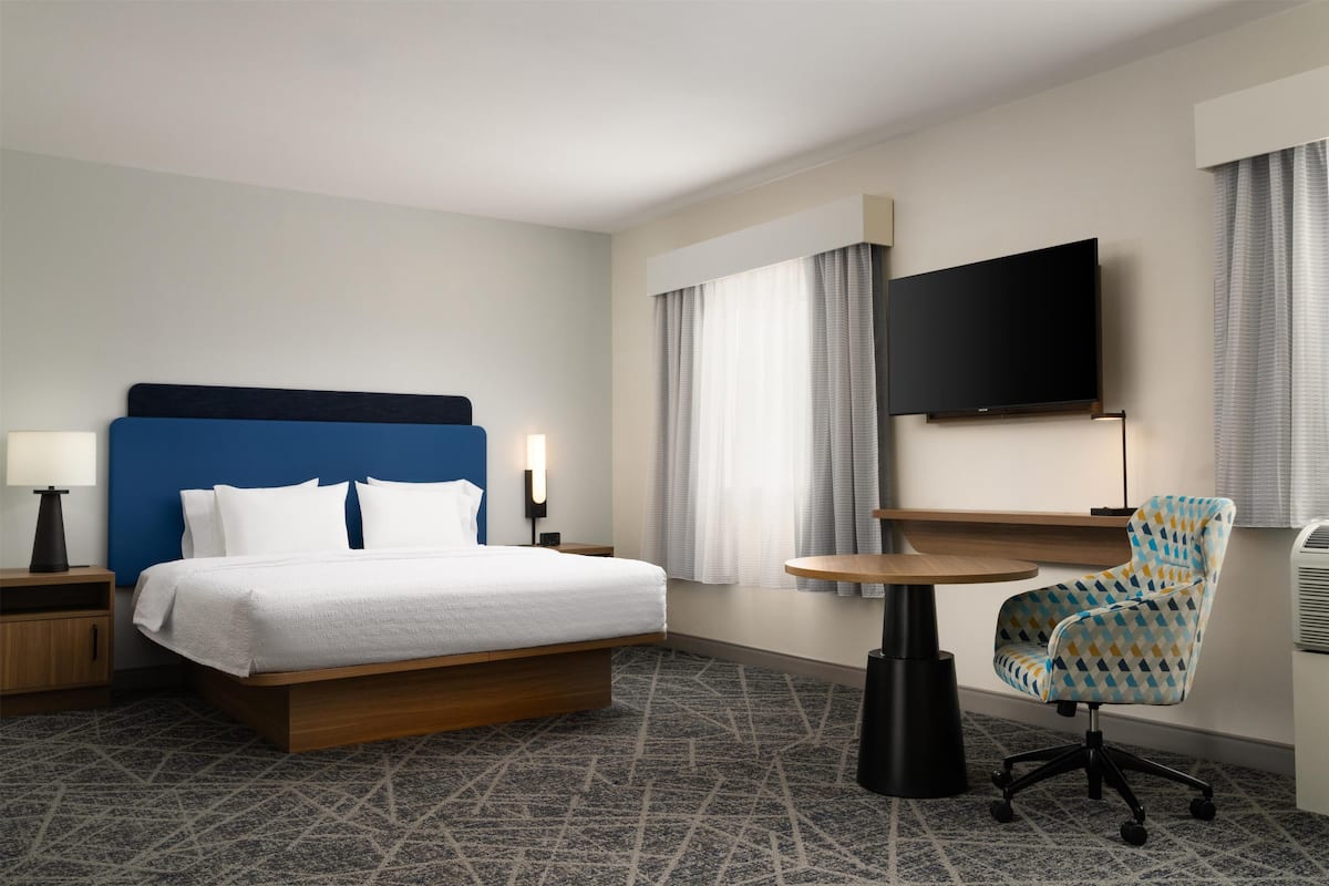 Habitación de hotel con cama, escritorio, silla, televisor y ventana con cortinas.