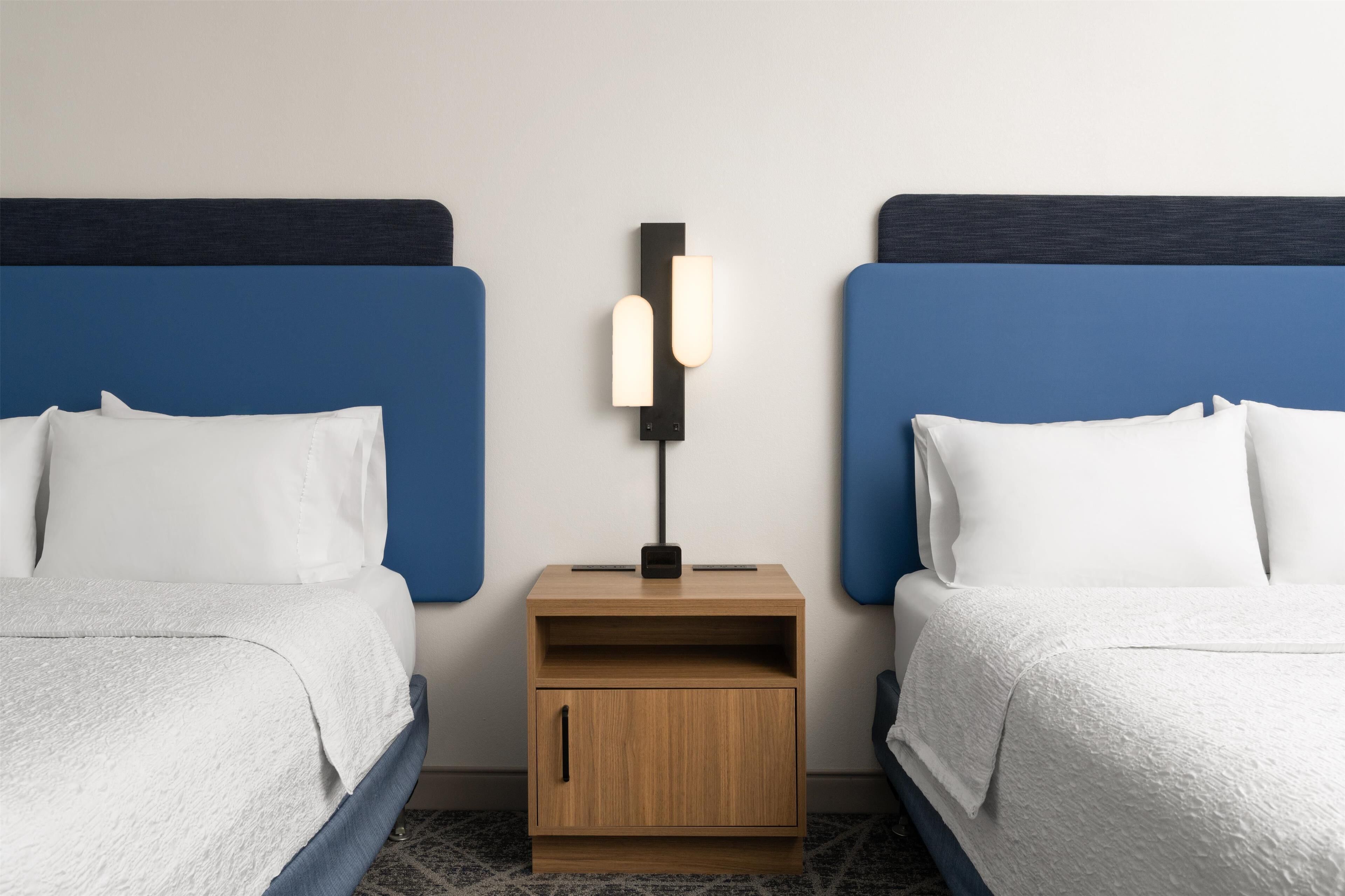 Una habitación de hotel con dos camas, una mesita de noche con lámpara y un pequeño armario de madera.