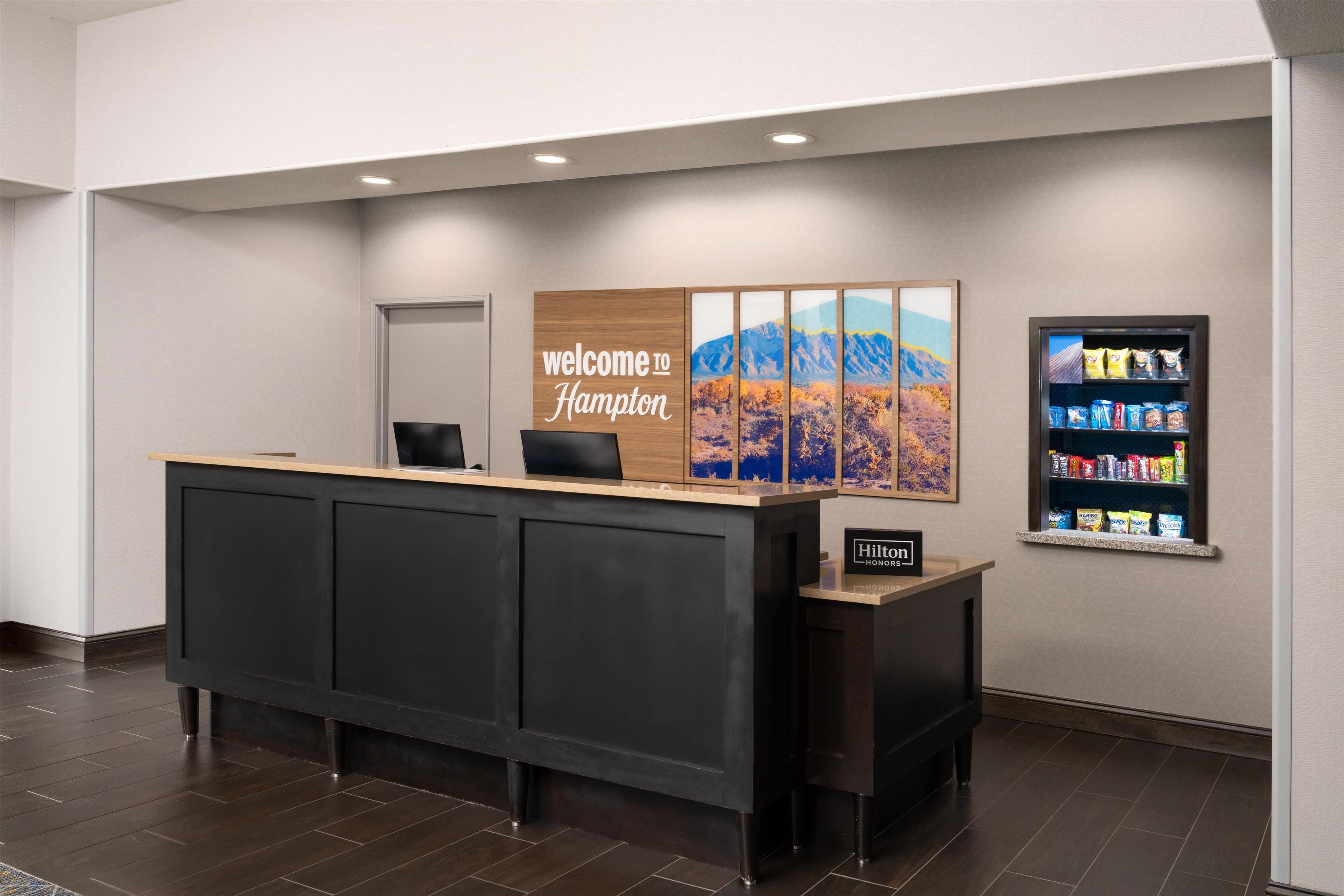 A reception desk with a 'Welcome to Hampton' sign, a framed landscape painting, and a snack vending machine.