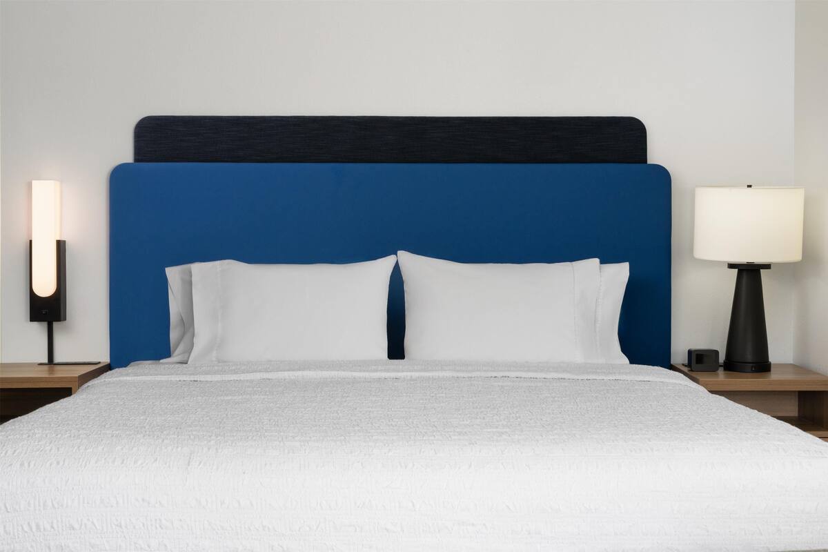 Una cama bien hecha con un cabecero azul, almohadas blancas y una colcha blanca. Hay dos mesitas de noche, cada una con una lámpara.
