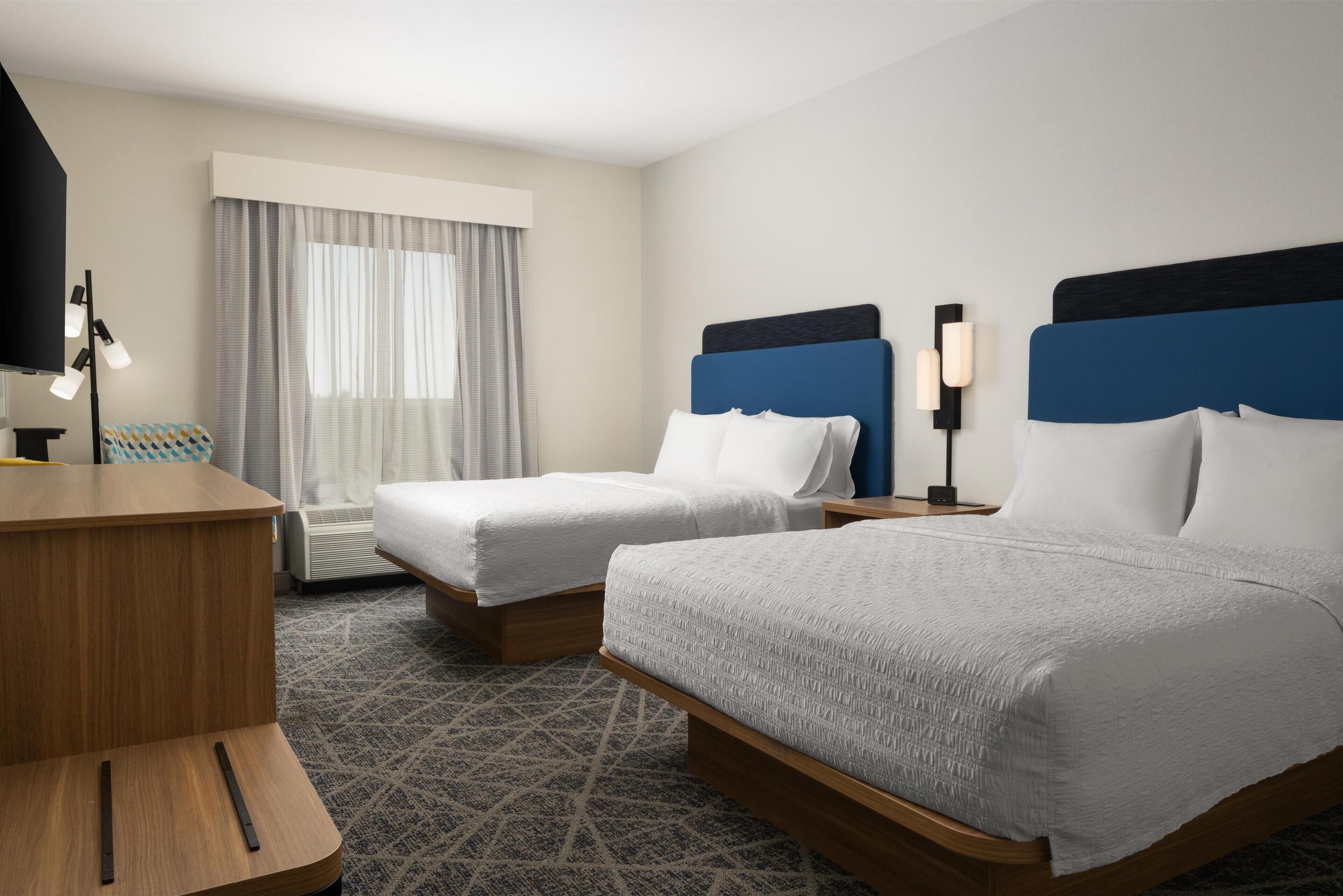 A hotel room with two beds, a desk, a lamp, and a window with curtains.