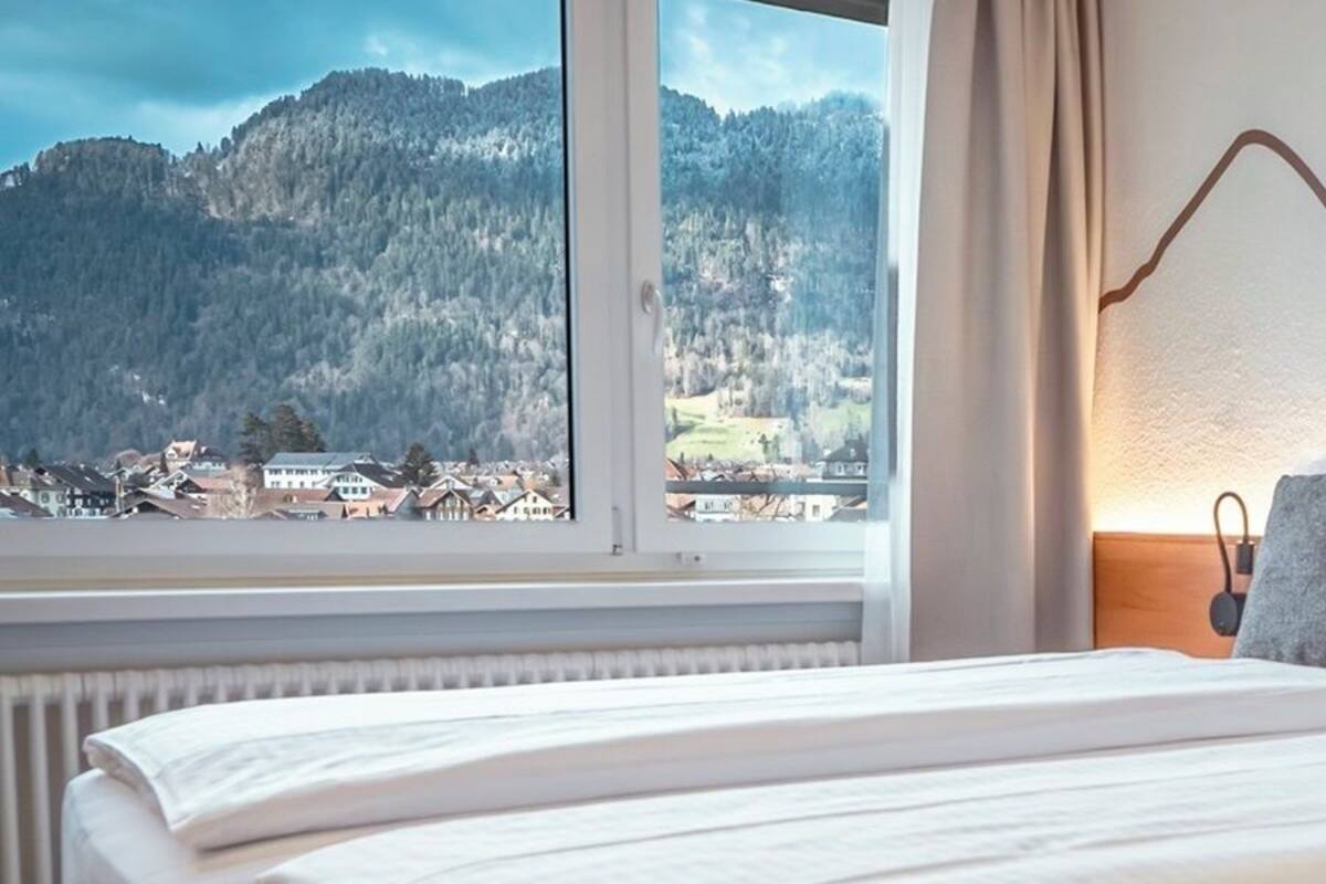 A hotel room with a large bed, a view of a mountainous landscape, and a wall-mounted light fixture.