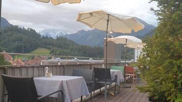 An outdoor terrace with tables and chairs, a view of mountains, and a tree.