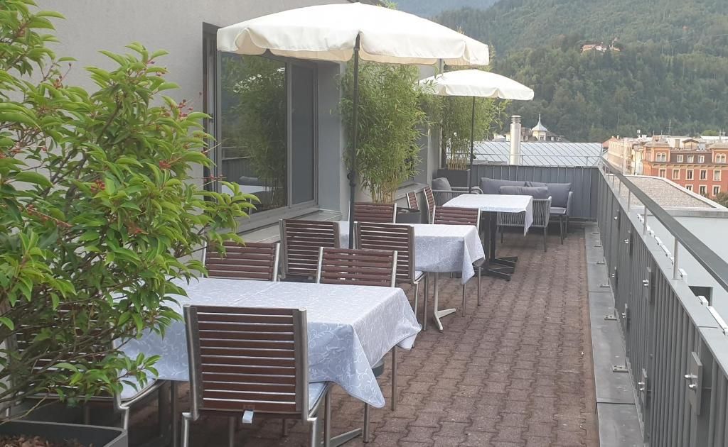 An outdoor terrace with tables and chairs, some covered by umbrellas, and a view of a cityscape and hills in the background.