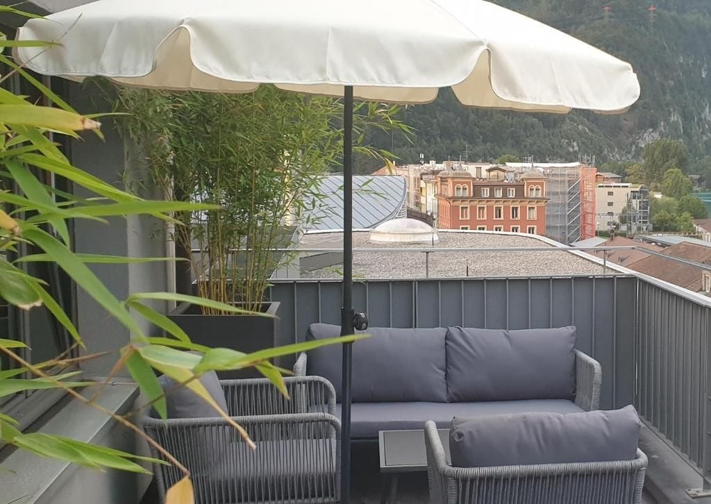 Ein Balkon mit einer grauen Couch und Sesseln, einem weißen Sonnenschirm und Blick auf Gebäude und Berge.
