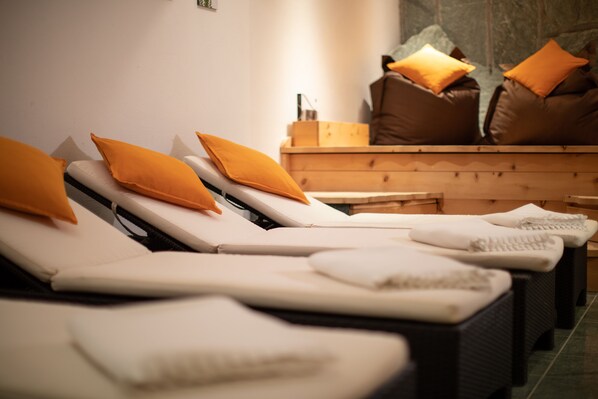 A row of sunbeds with orange cushions, a wooden bench, and a stone wall in the background.