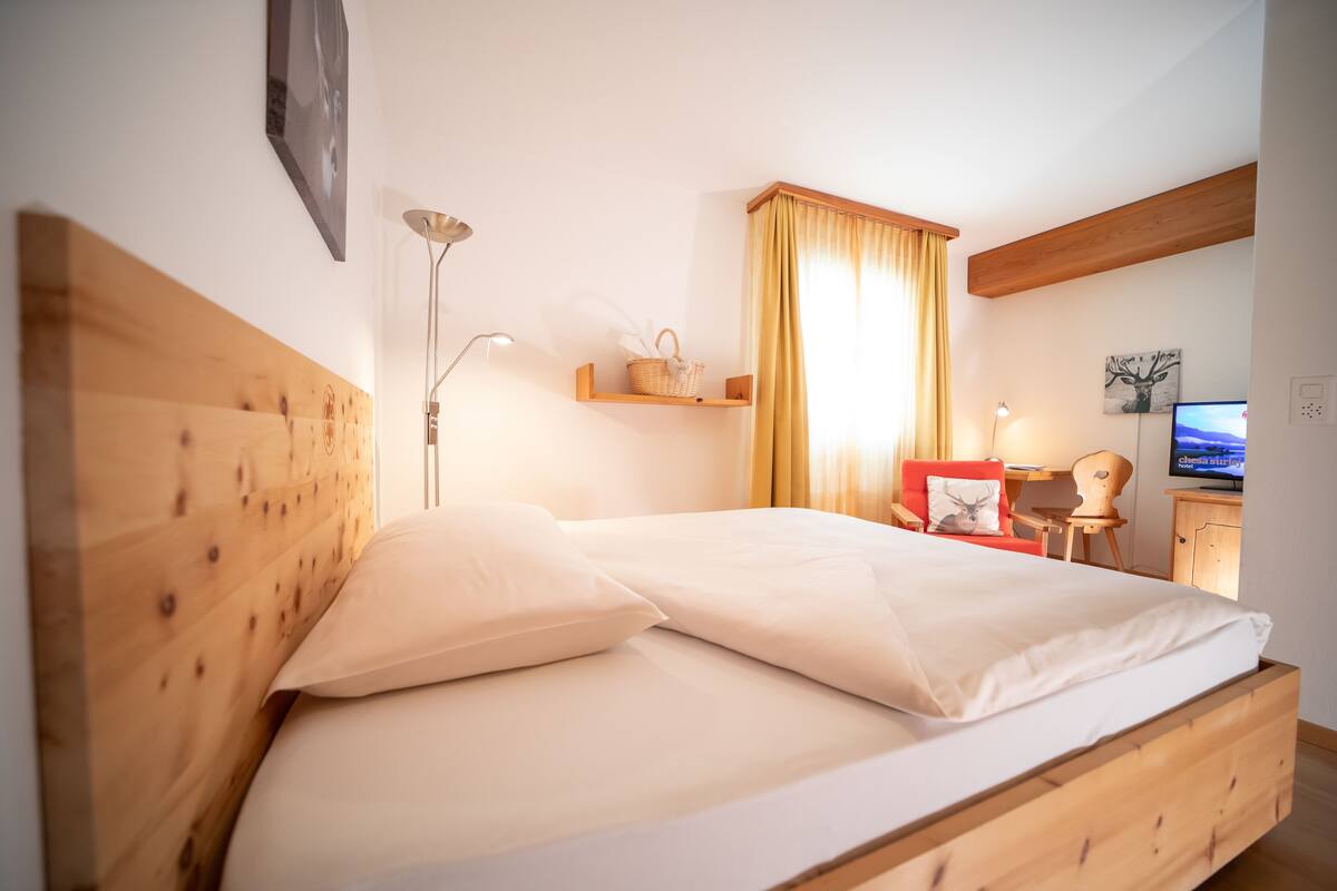 A wooden bed with white linens, a bedside lamp, a shelf with a basket, a desk with a chair, and a television.