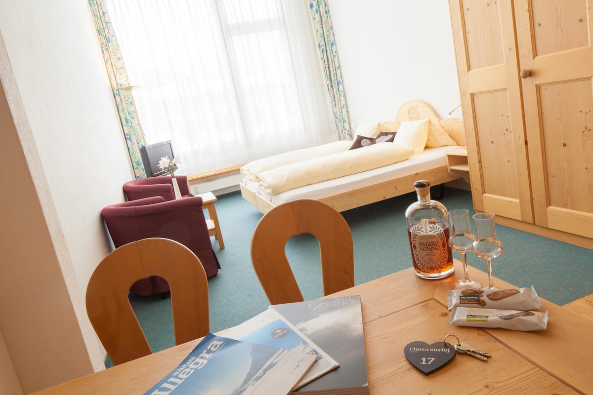 A hotel room with a bed, a wooden table with a magazine, a bottle of liquor, and glasses.