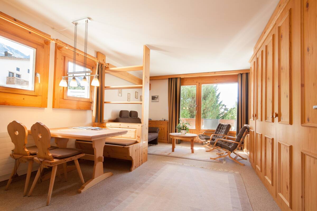 A wooden interior with a dining table, chairs, a sofa, and a rocking chair. There are windows providing natural light and a view of the outdoors.